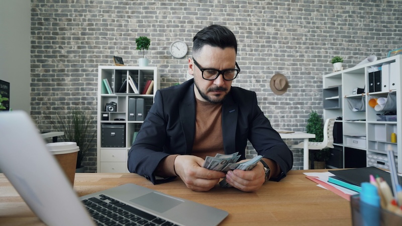 man anxiously counting money at a desk near an open computer