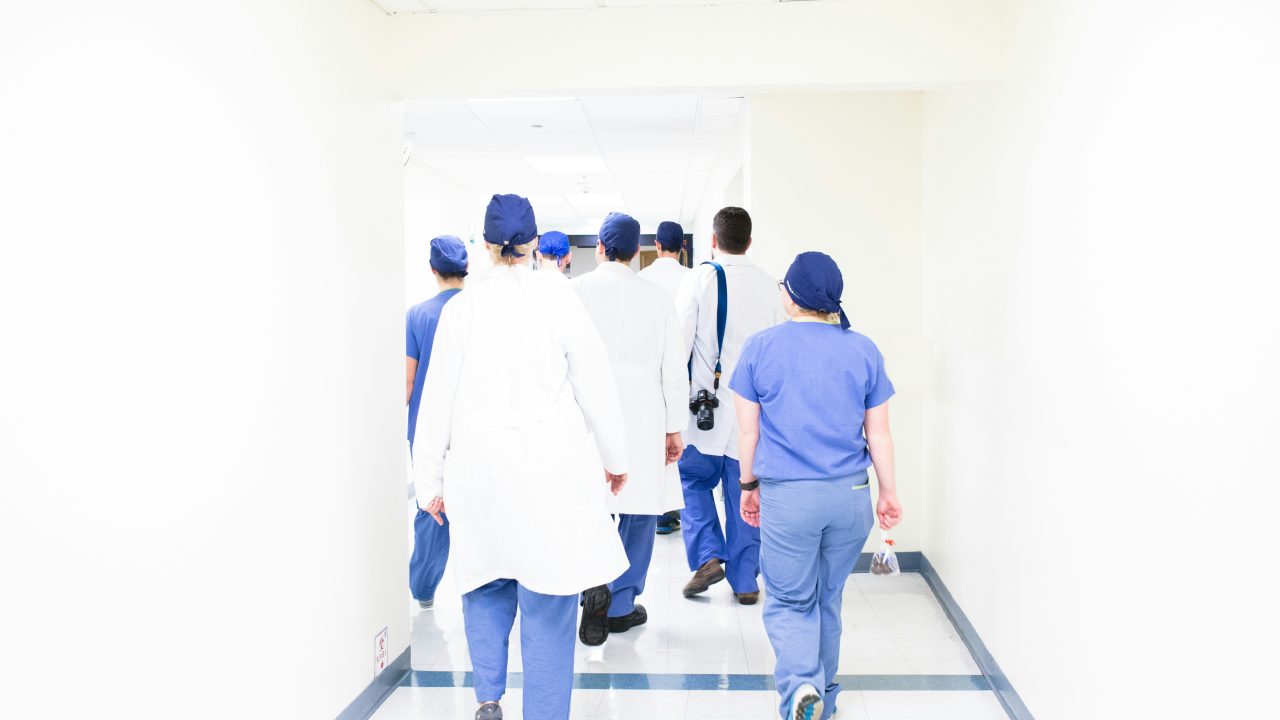 white hospital hallway as doctors and nurses in labcoats and scrubs walk down a cooridor