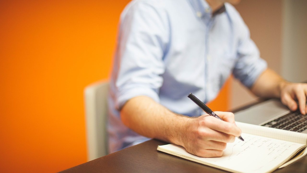 man writing on notepad at desk with a bright orange background
