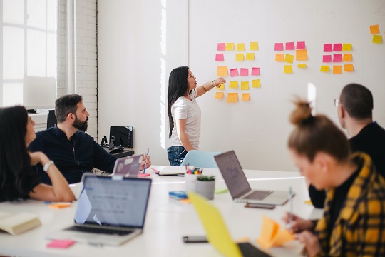 group of employees sitting around a large table with their laptops open while one person stands at the whiteboard putting sticky notes on the wall