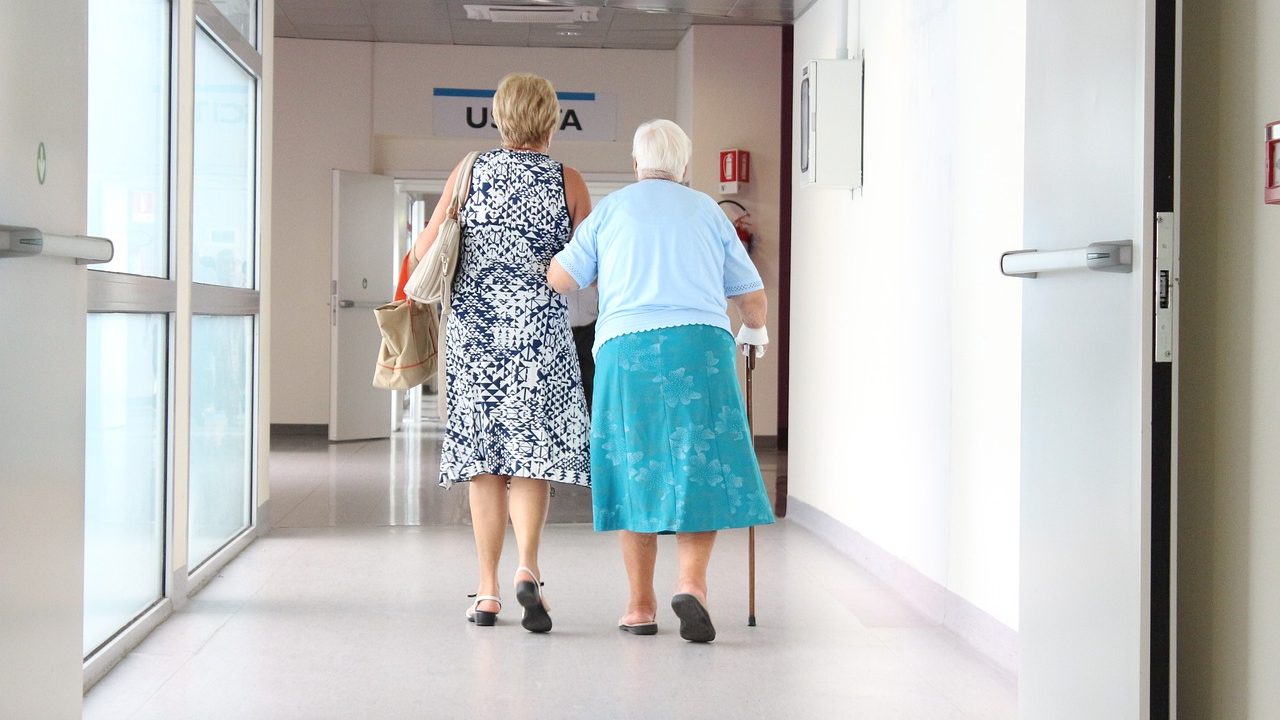 elderly woman walking elbow to elbow with a younger woman down an aisle of a medical building